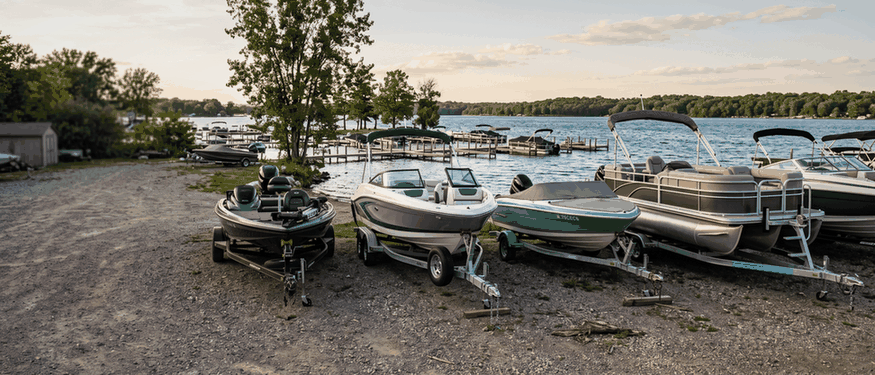Pontoon, bass and bowrider boats on trailers at a lakeside boat dealer, each priced differently to insure